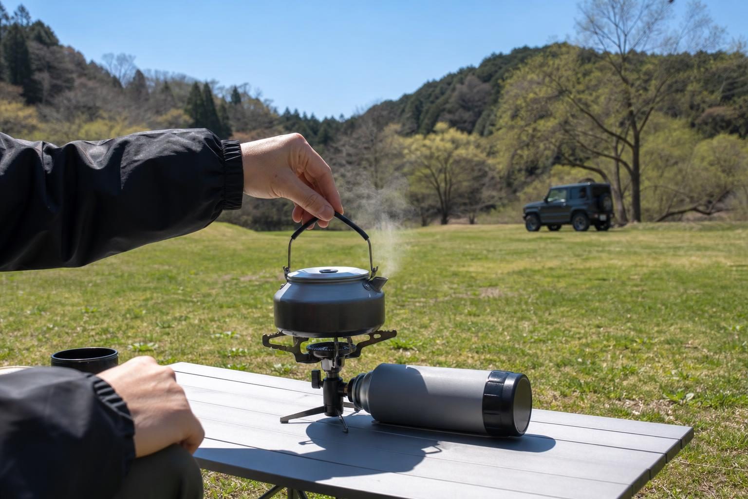 晴れた日中の芝生の上。車から離れた場所の低いテーブルに置いた小型のシングルバーナーでお湯を沸かしている様子