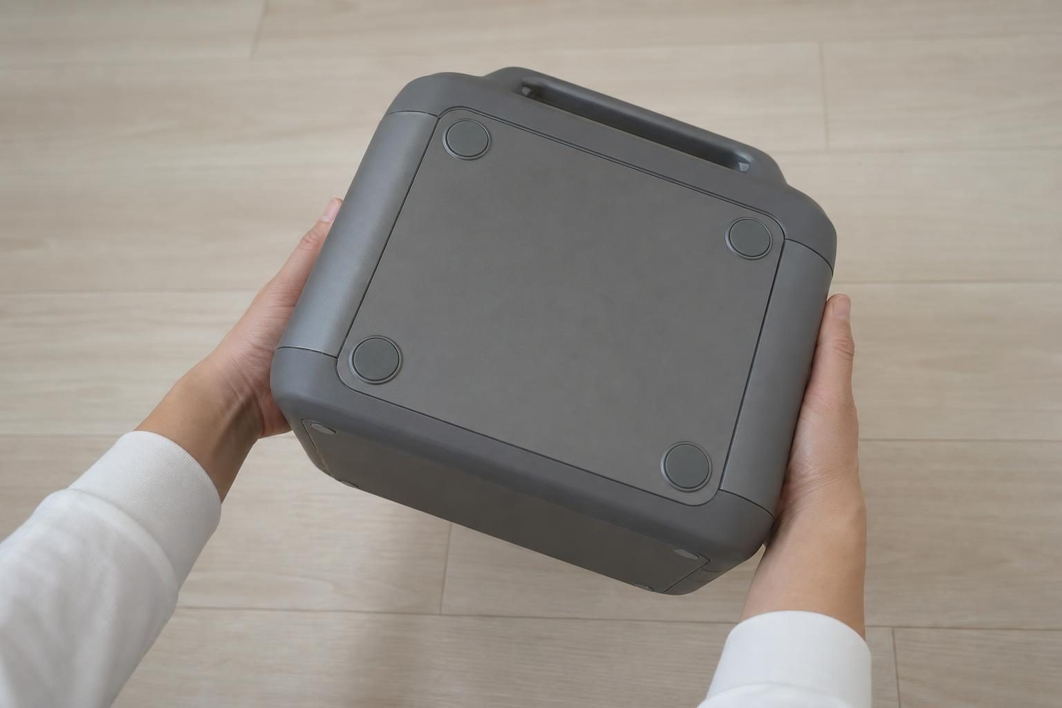 屋内の床に置かれた、ロゴや文字のない無地の箱型ポータブル電源を、手元だけ写した人が裏返して底面を見ている様子。キャンプ道具は写さない