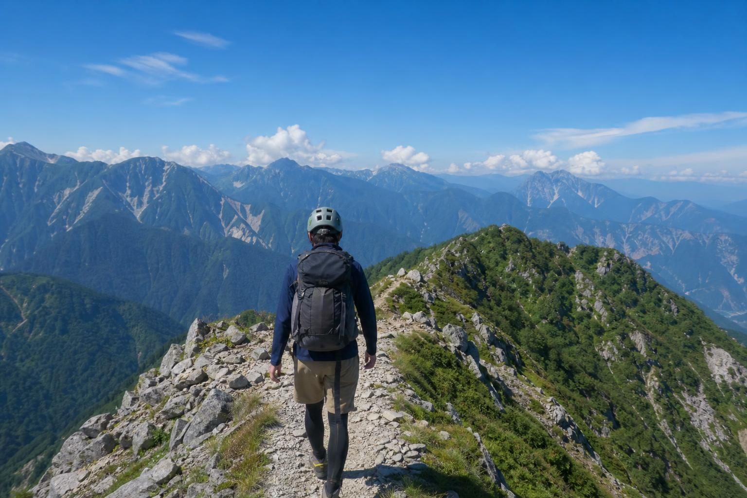 岩の多い稜線の登山道を、ヘルメットをかぶりザックを背負って歩く登山者の後ろ姿