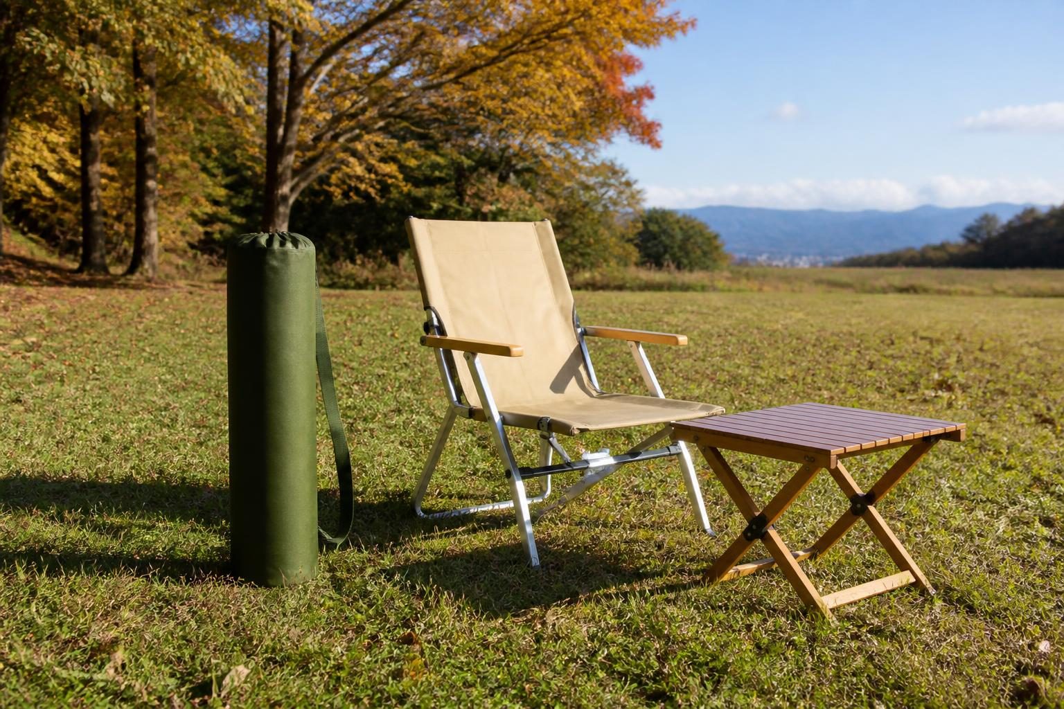 紅葉の草地に立てた無地のオリーブ色の円筒形バッグ(肩ひも付き)と、折りたたみチェア・木のテーブル