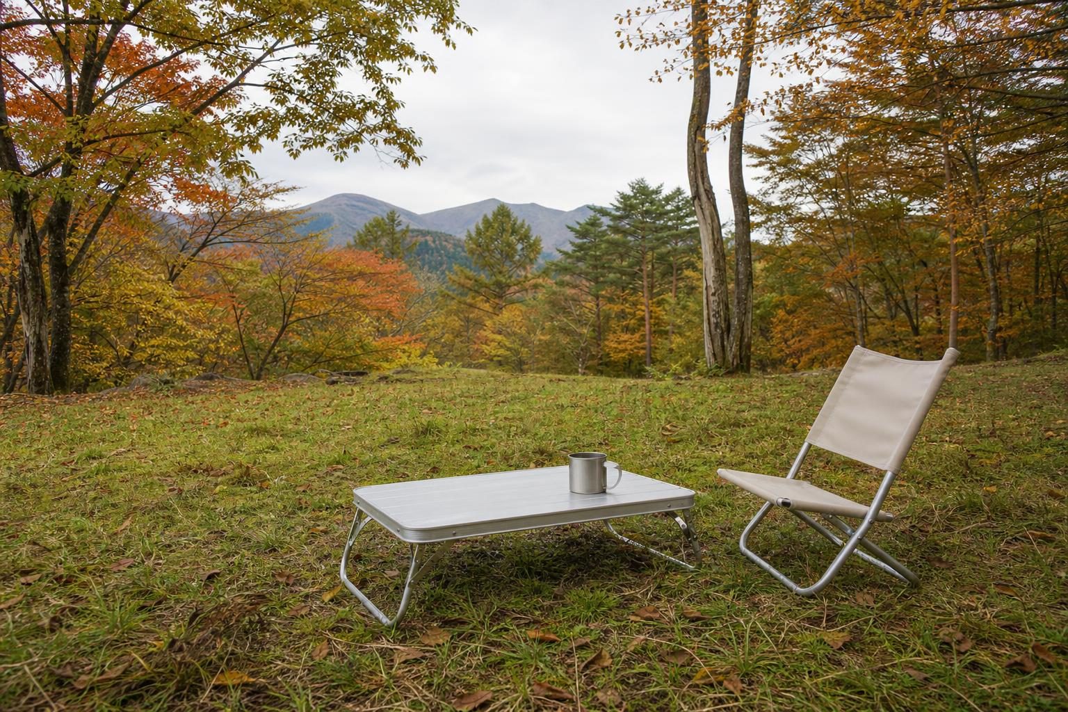 紅葉した山あいの芝生に置いた、低いアルミのテーブルとステンレスのマグ、ベージュの折りたたみチェア